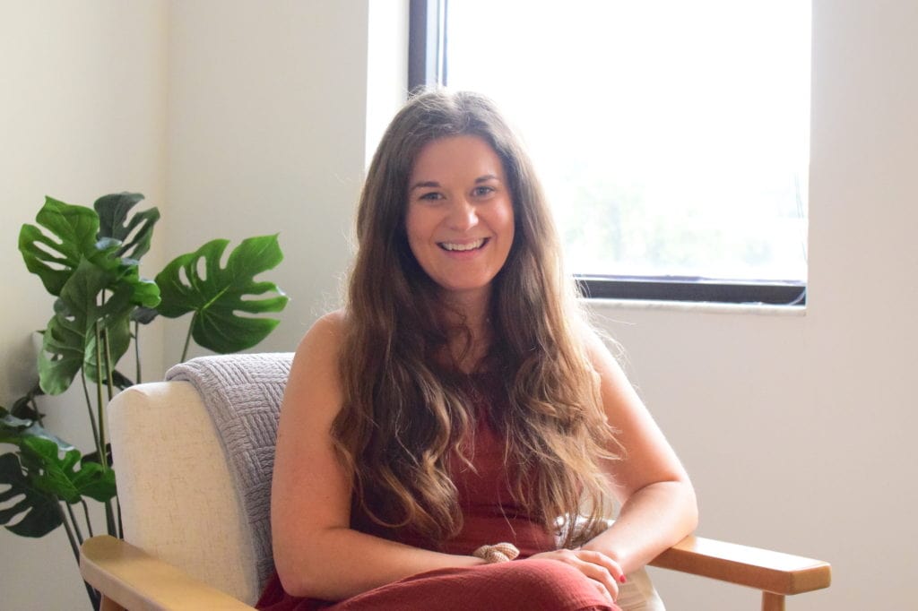 Emily Murray, a woman in a rust-red sundress with long brown hair, sits in an armchair in her office at Murray Nutrition.