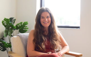 Emily Murray, a woman in a rust-red sundress with long brown hair, sits in an armchair in her office at Murray Nutrition.