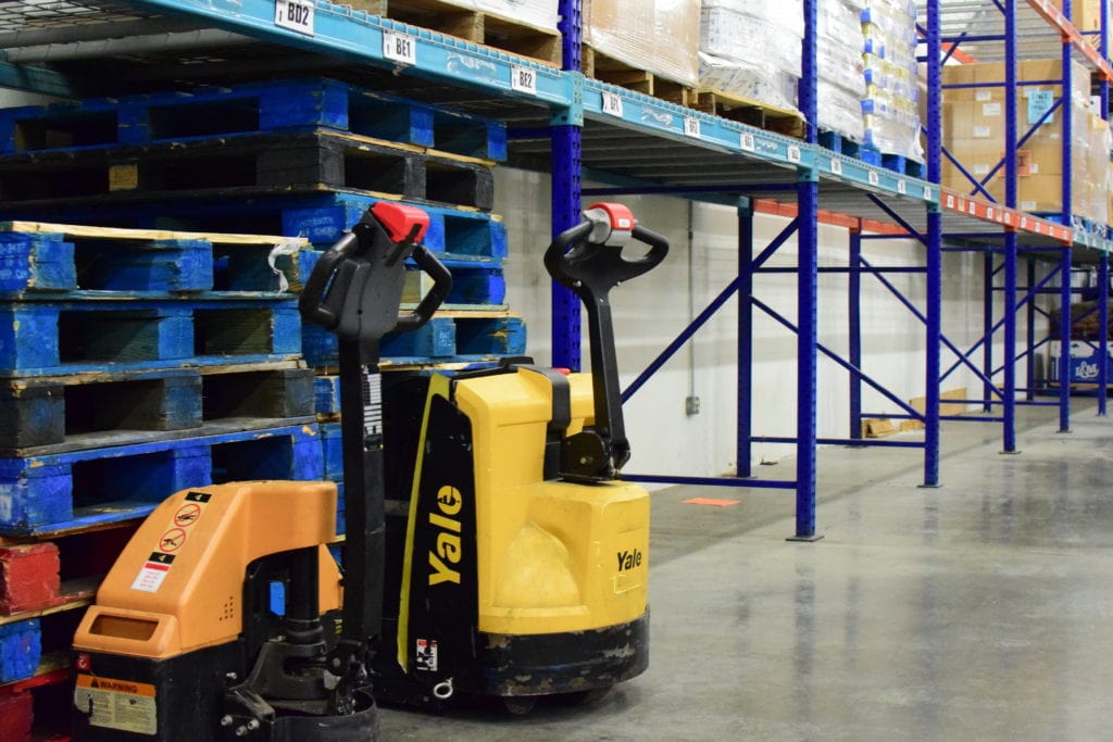 Two pallet jacks sit beside empty blue wooden pallets, and shelves along a wall in the OneGenAway warehouse sit empty.
