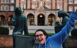 Adrienne Pelletier, a woman in a blue hoodie, strikes a pose in front of the Notre Dame Seminary building.