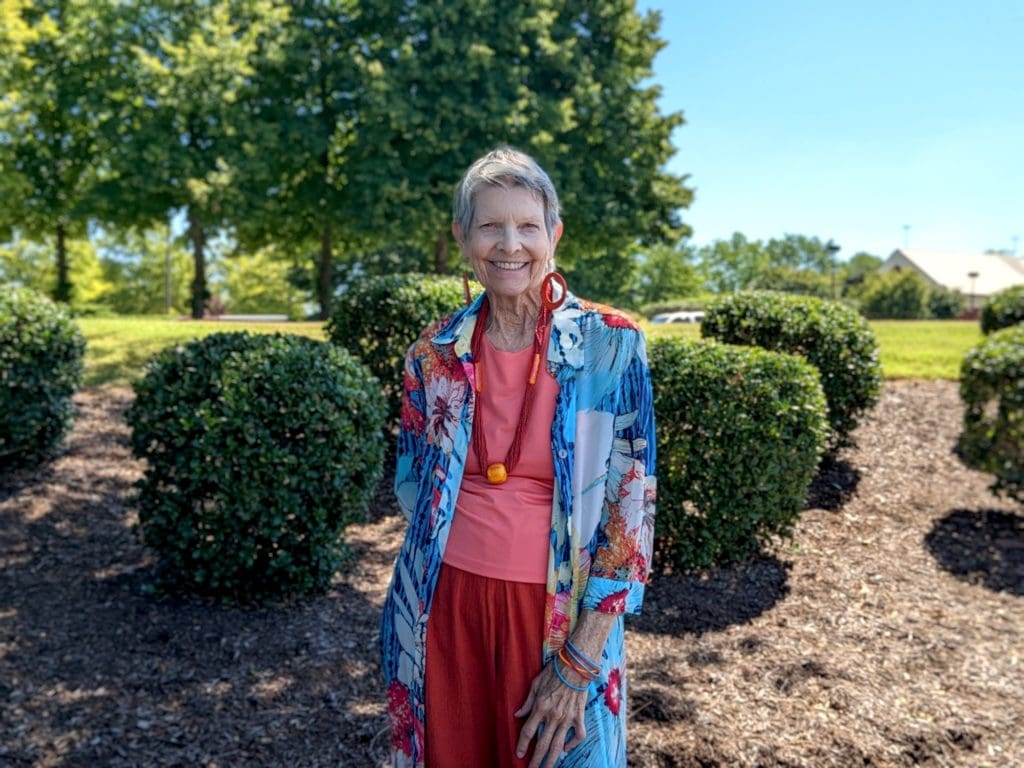 A woman in a pink shirt, red pants, and blue patterned shawl stands in front of trees in bushes