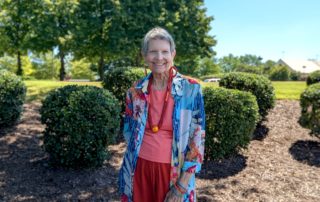A woman in a pink shirt, red pants, and blue patterned shawl stands in front of trees in bushes